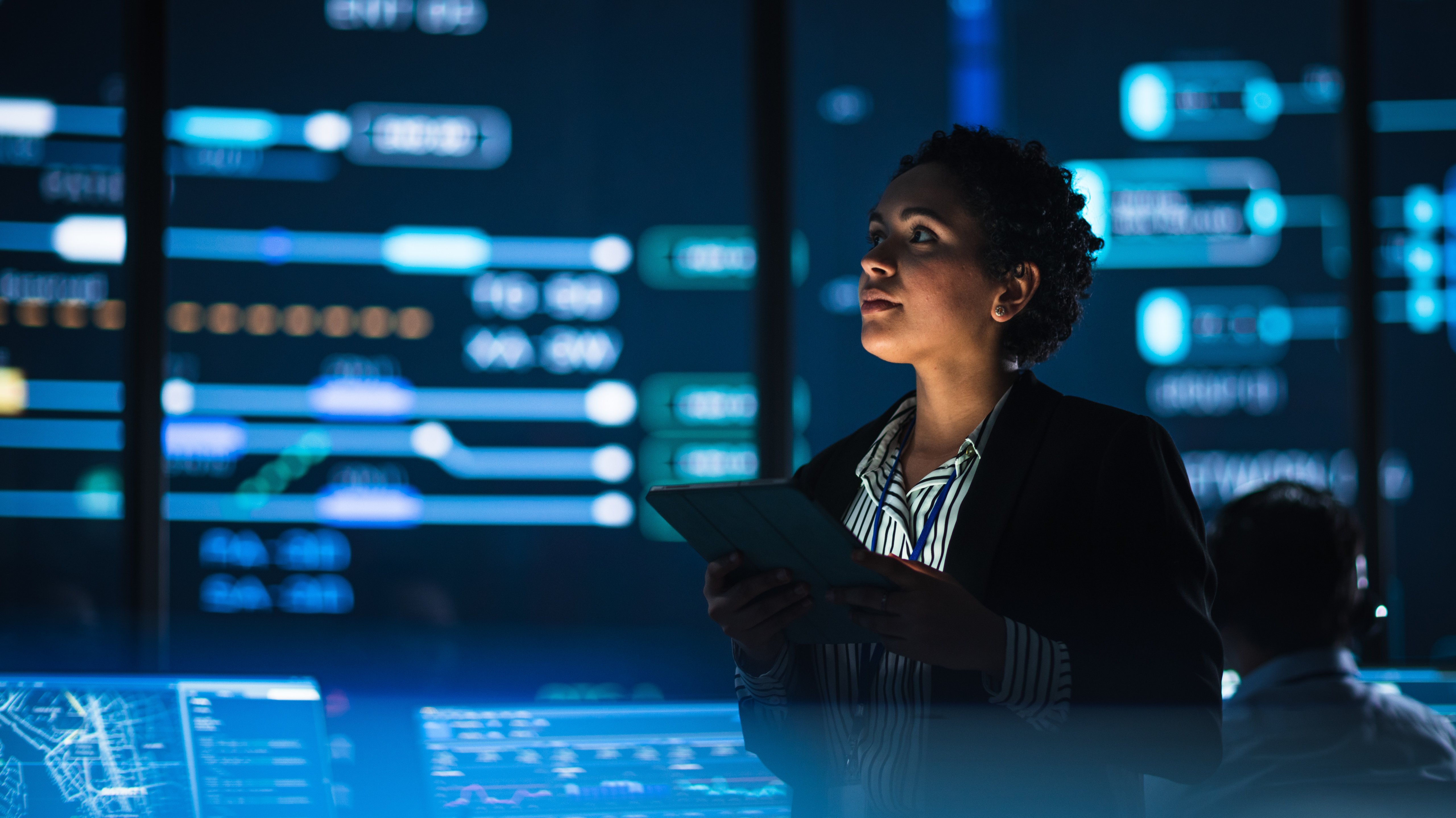 Young Multiethnic Female Government Employee Uses Tablet Computer in System Control Monitoring Center. In the Background Her Coworkers at Their Workspaces with Many Displays Showing Technical Data.